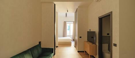 A narrow hallway with a green sofa on the left, a wooden cabinet with a television on the right, and a bed at the end of the hallway.