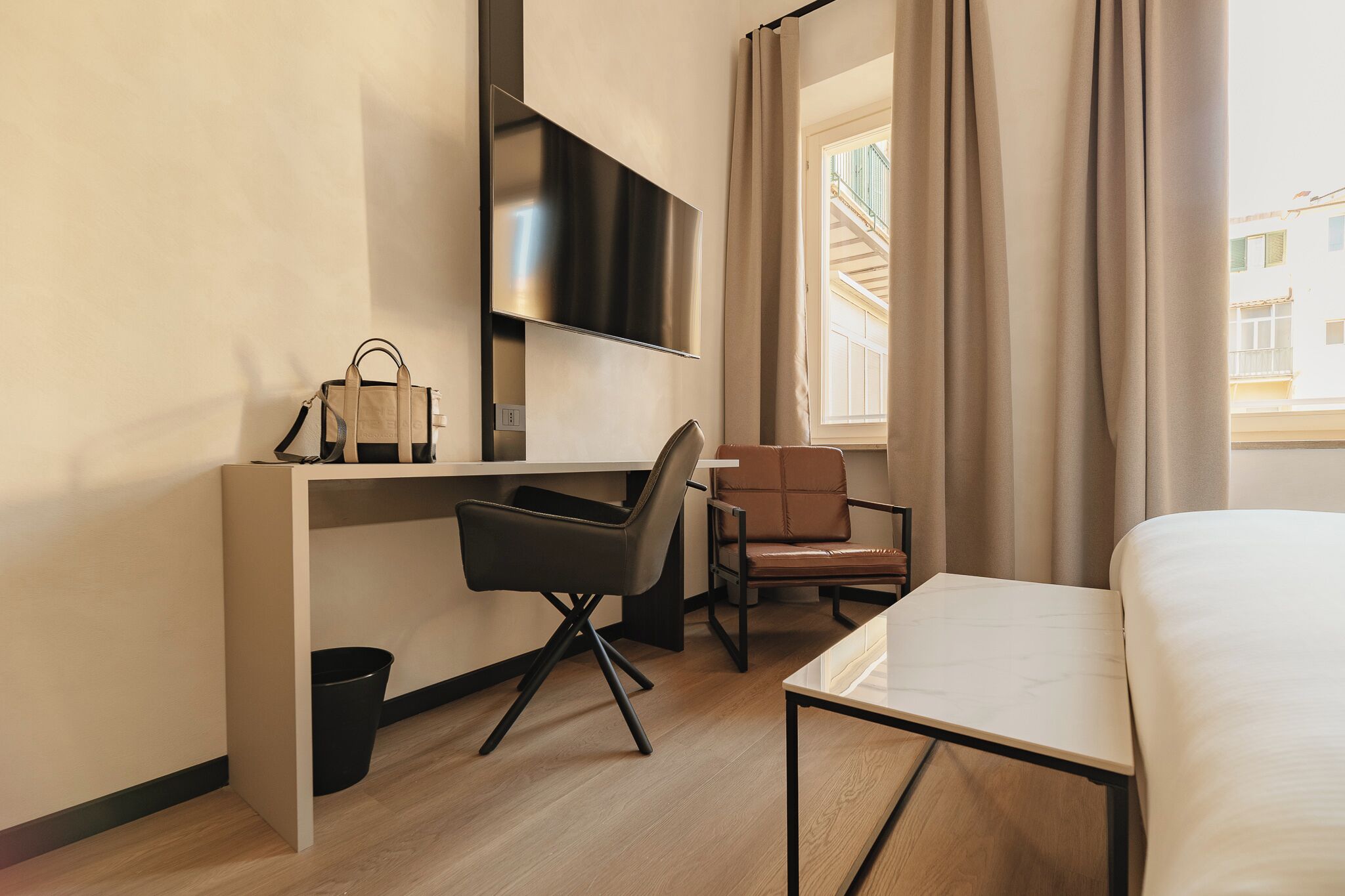 A hotel room with a bed, a desk with a chair, a TV mounted on the wall, and a window with curtains.