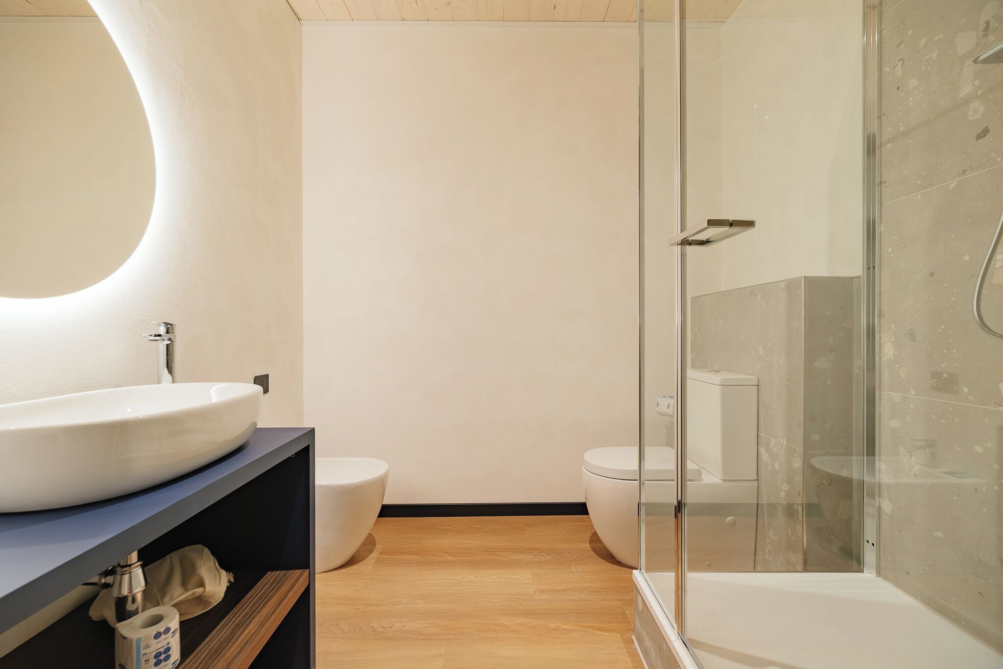 A modern bathroom with a large oval mirror, a white sink, two toilets, and a glass-enclosed shower area.