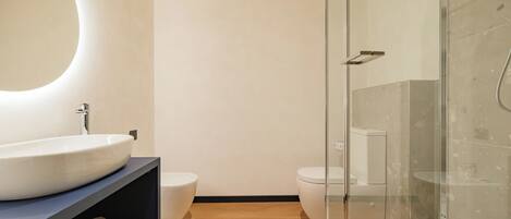 A modern bathroom with a large oval mirror, a white sink, two toilets, and a glass-enclosed shower area.