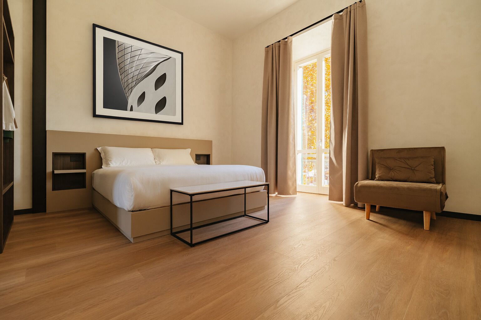 A modern bedroom with a bed, bedside tables, a bench, a chair, and a large window with curtains.