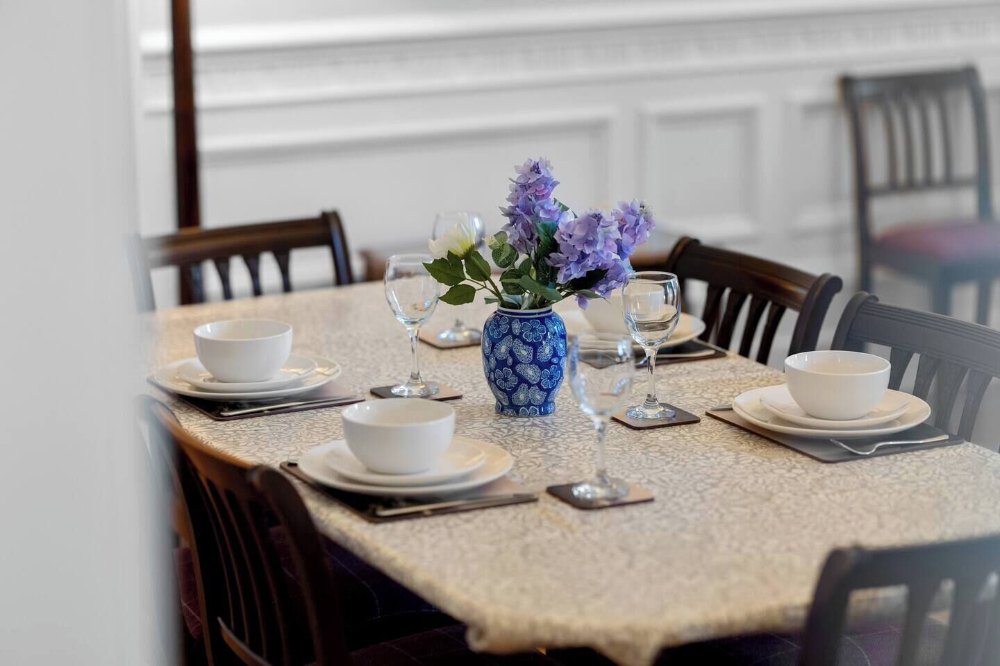 A dining table set with white bowls, plates, and cutlery, a blue vase with purple flowers, and a textured tablecloth.