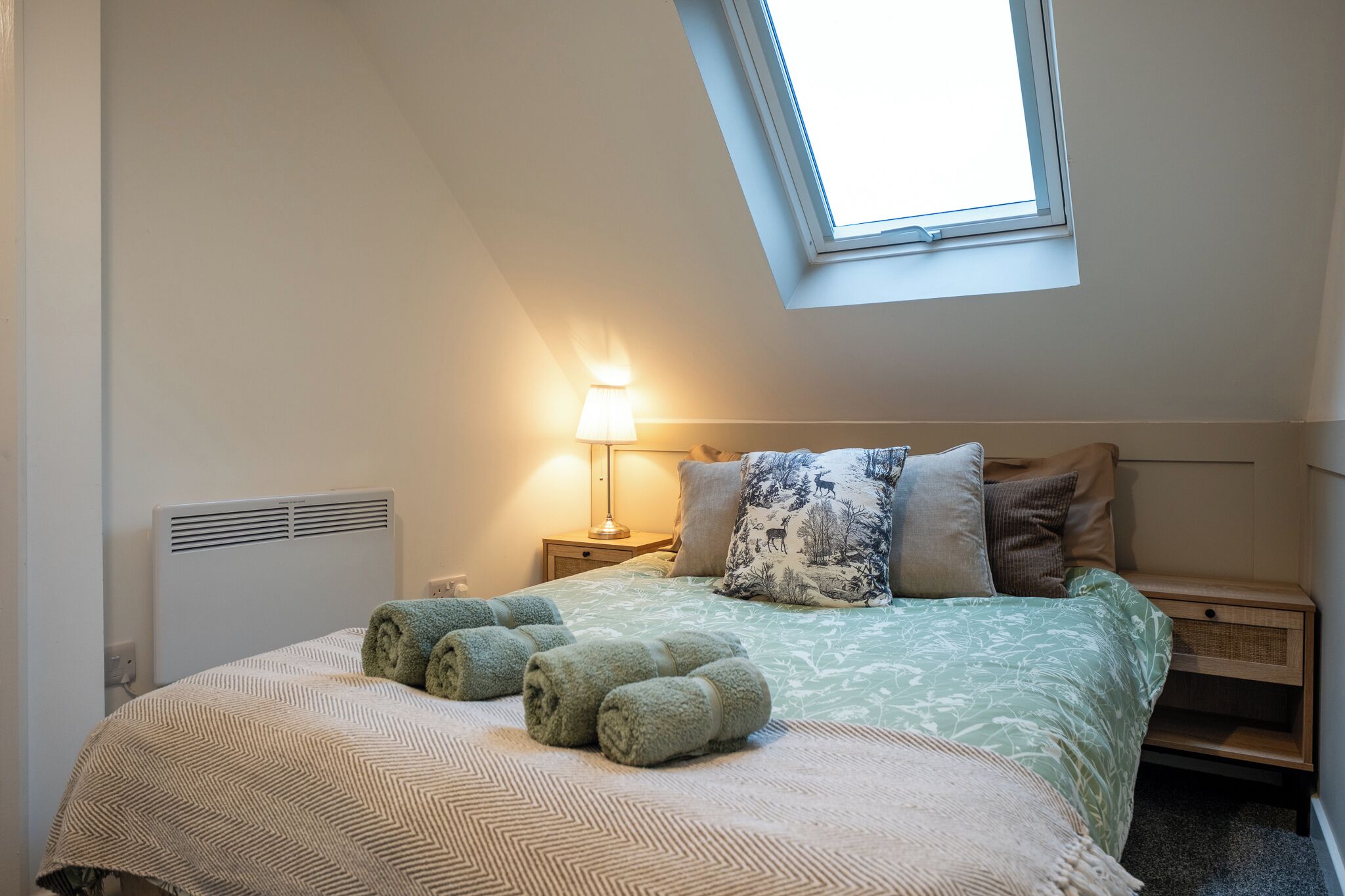A bedroom with a skylight, a bed with rolled towels, a bedside lamp, and a nightstand.