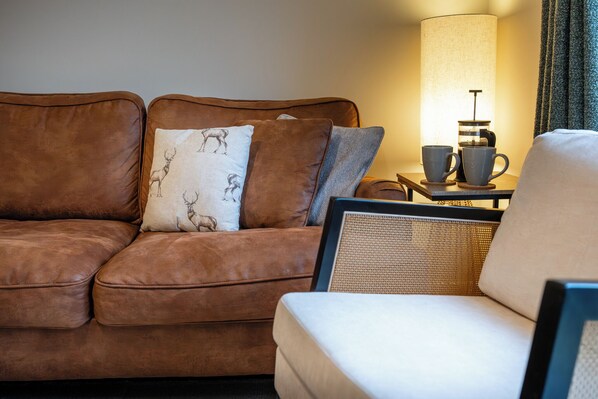 Een bruine leren bank met gekleurde kussens, een witte fauteuil, een bijzettafeltje met een lamp, en twee mokken.