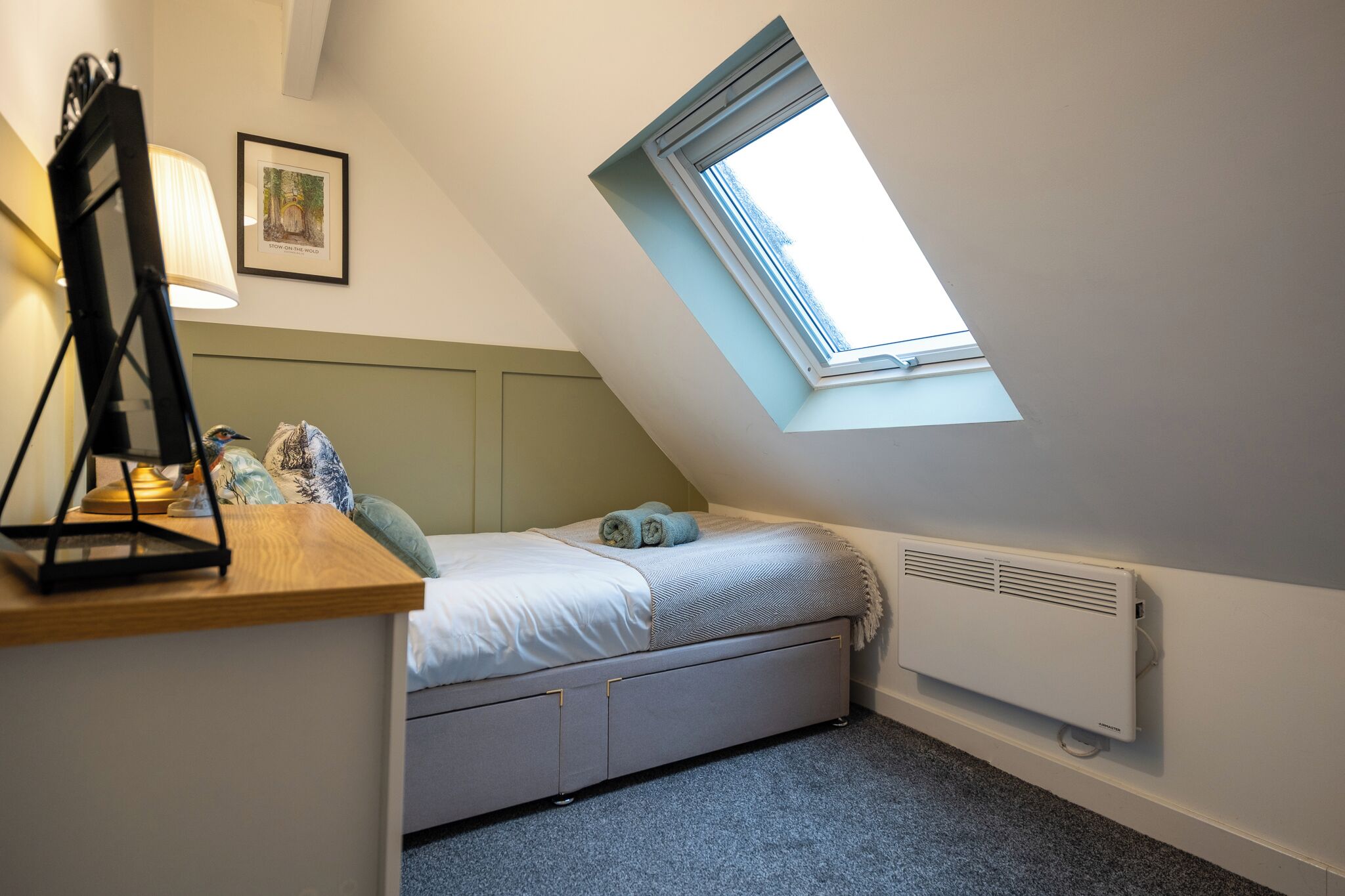A small, cozy bedroom with a sloped ceiling, a skylight, a bed with a headboard, a bedside table with a lamp, and a radiator.