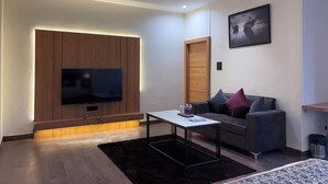 Un salon moderne avec un téléviseur à écran plat fixé sur un mur recouvert de panneaux en bois, un canapé gris avec des coussins, une petite table blanche et un tapis noir.