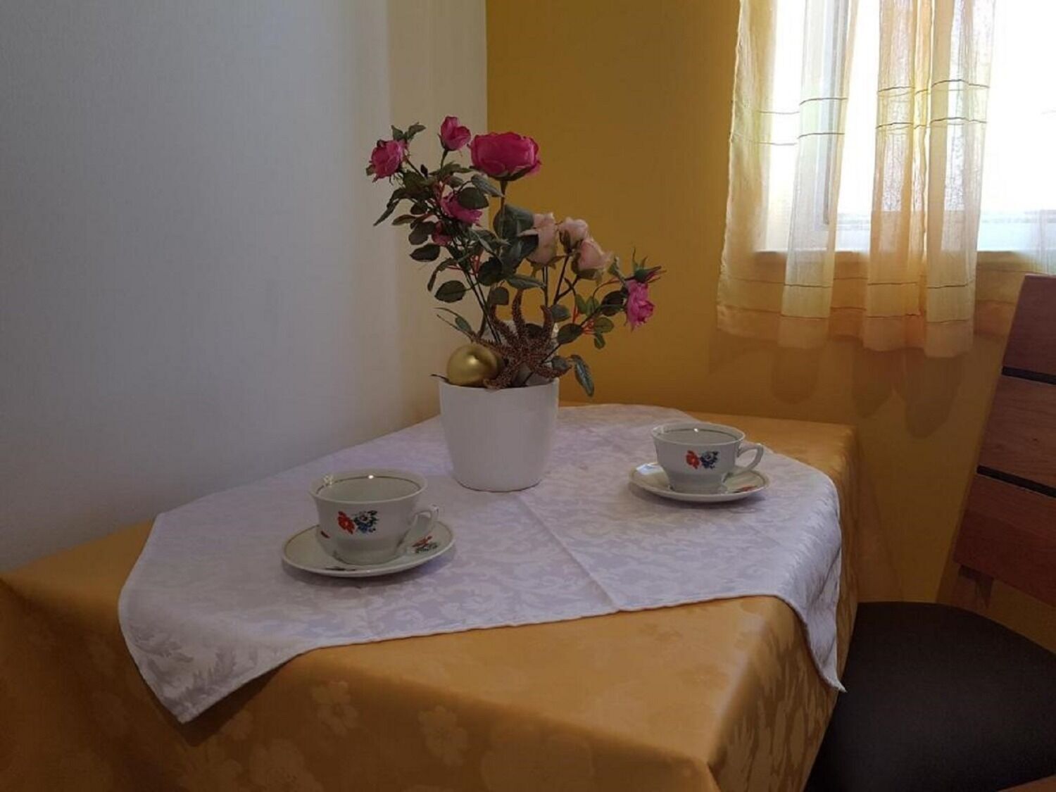 Une petite table recouverte d’une nappe blanche, deux tasses blanches ornées de motifs floraux et un pichet blanc décoré de roses roses.