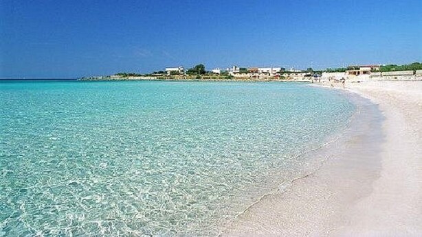 A beach with clear turquoise water, a sandy shore, and buildings in the background. - Apartment 'Monolocale Eddy By Salentolimit' with Balcony and Air Conditioning (Torre Lapillo)
