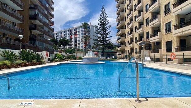 En swimmingpool omgiven av höga byggnader med balkonger, en klar himmel och en livboj vid poolens kant.