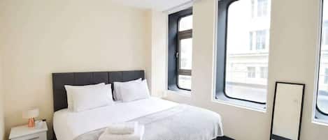 Habitación de hotel con cama, mesita de noche, espejo y ventana con vista a edificios.