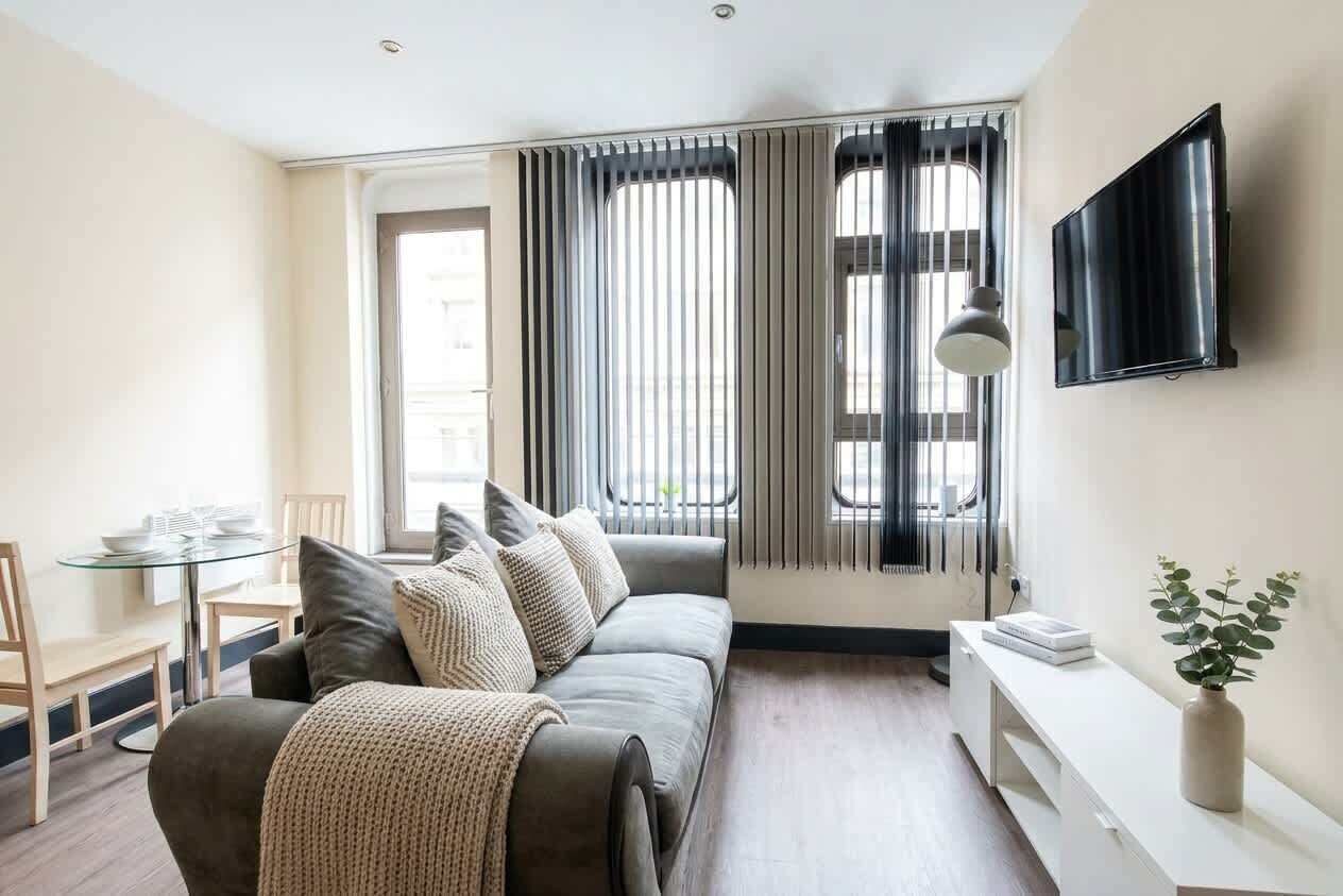 Een moderne woonkamer met een bank, een glazen tafel, een aan de muur bevestigde televisie en een raam met jaloezieën.