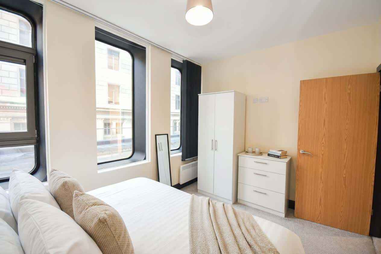 Een slaapkamer met een bed, een kledingkast, een dressoir en een raam met uitzicht op gebouwen.