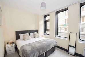 Ein Hotelzimmer mit Bett, Nachttisch, Lampe und einem Fenster mit Blick auf Gebäude.