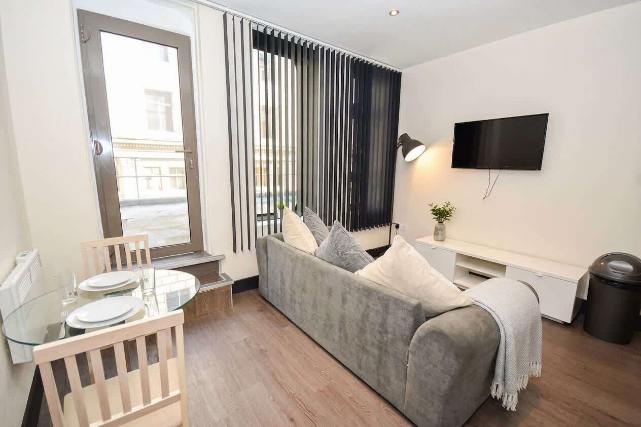 Un salon moderne avec un canapé gris, une table à manger en verre et un téléviseur fixé au mur.