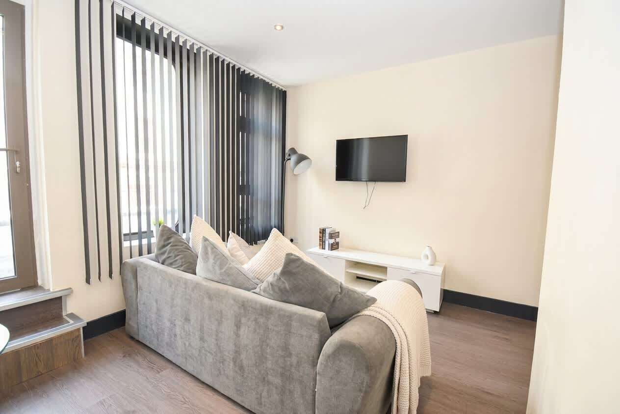 A modern living room with a grey sofa, a flat-screen TV mounted on the wall, and a white side table.