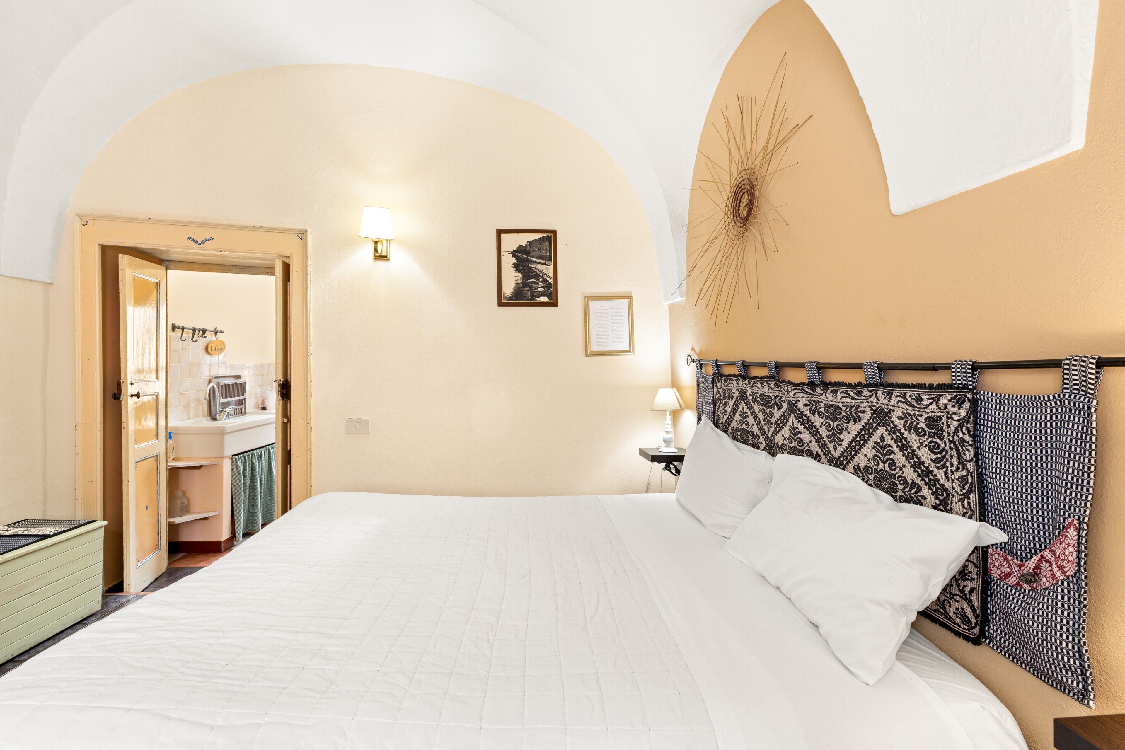A bedroom with a bed, a nightstand, a wall-mounted lamp, a framed picture, and an arched doorway leading to a bathroom.