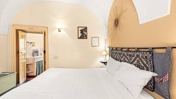 A bedroom with a bed, a nightstand, a wall-mounted lamp, a framed picture, and an arched doorway leading to a bathroom.