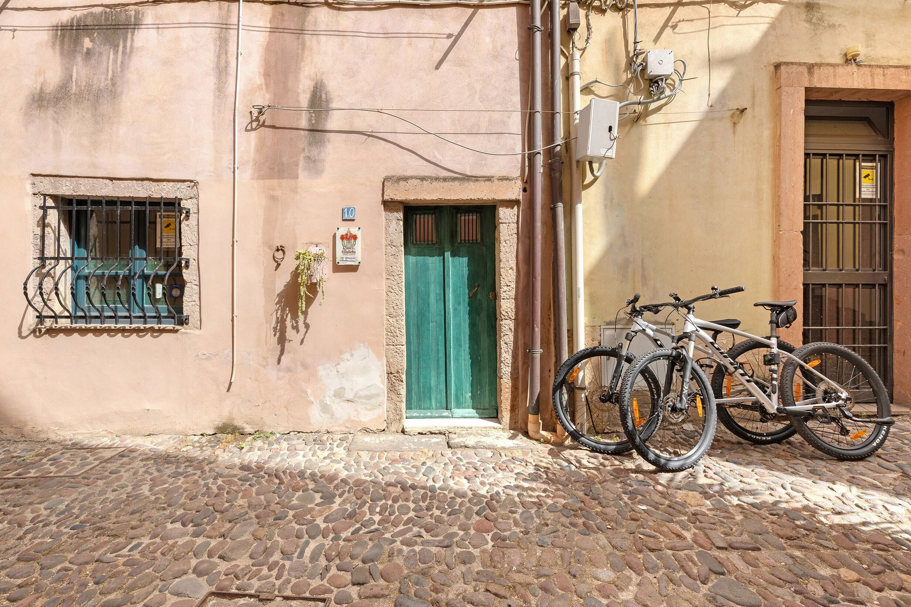 Une rue pavée, avec un bâtiment dont la porte est peinte en vert, une fenêtre ornée de barreaux en métal et un vélo garé à l’extérieur.