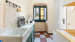Une petite cuisine équipée d’un évier, d’une fenêtre avec volets et d’une porte sur laquelle est apposée une enseigne.