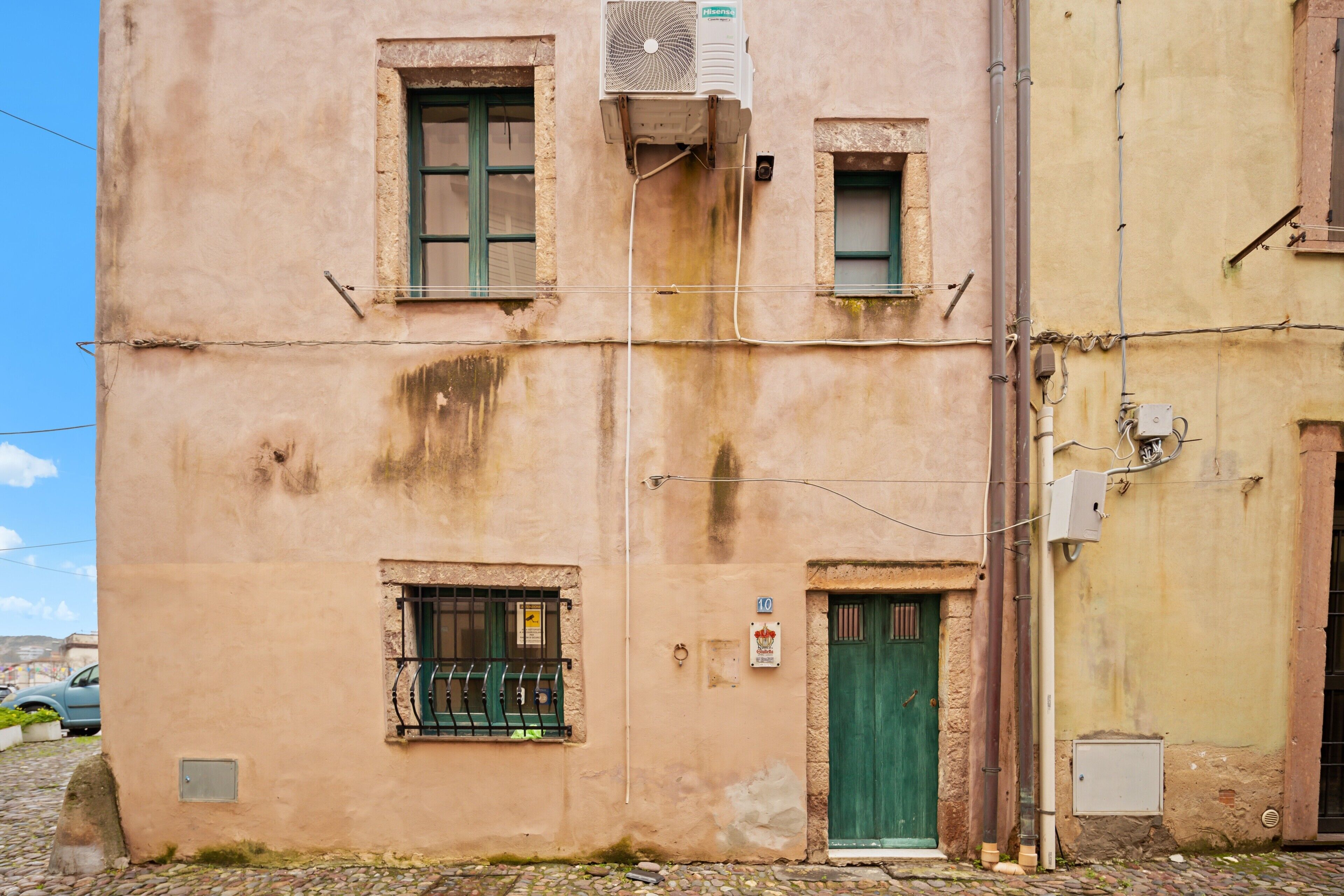 A building with a green door, a window with a metal grill, and an air conditioning unit mounted on the wall.