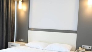 A neatly made bed with white linens and a folded towel, a bedside table with a phone, and a wall-mounted light fixture. - HOTEL ROXX ROYAL (Kusadasi)