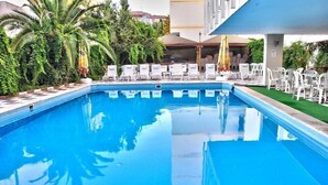 A swimming pool with white lounge chairs and umbrellas, surrounded by greenery and a building in the background. - HOTEL ROXX ROYAL (Kusadasi)