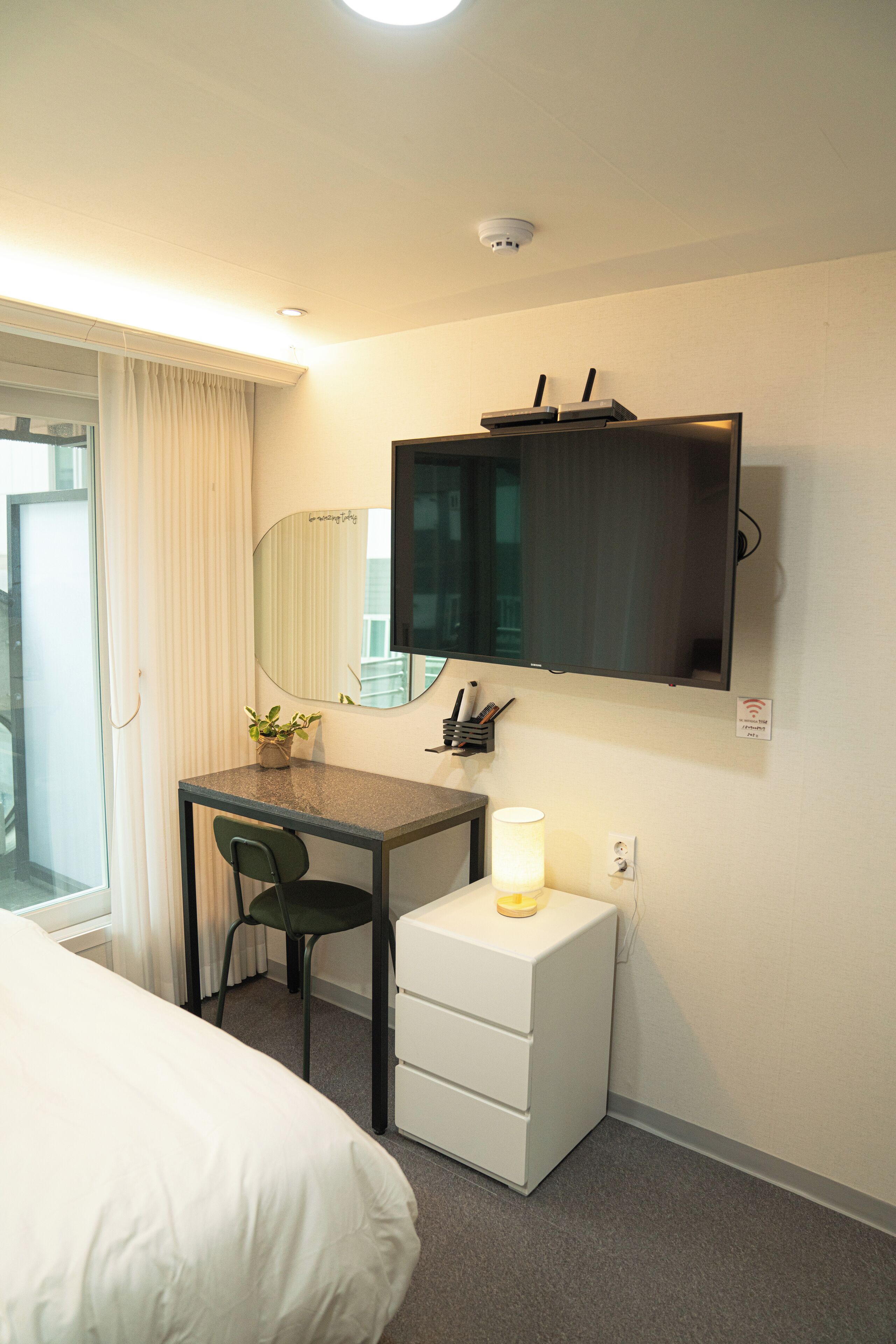 Ein Hotelzimmer mit einem Bett, einem Schreibtisch mit Stuhl, einem kleinen weißen Nachttisch, einem an der Wand befestigten Fernseher, einem Spiegel und einem Fenster mit Vorhängen.
