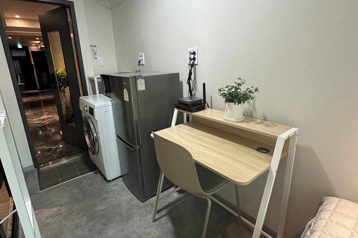 A compact room with a bed, a desk with a chair, a washing machine, a refrigerator, and an air conditioner mounted on the wall.