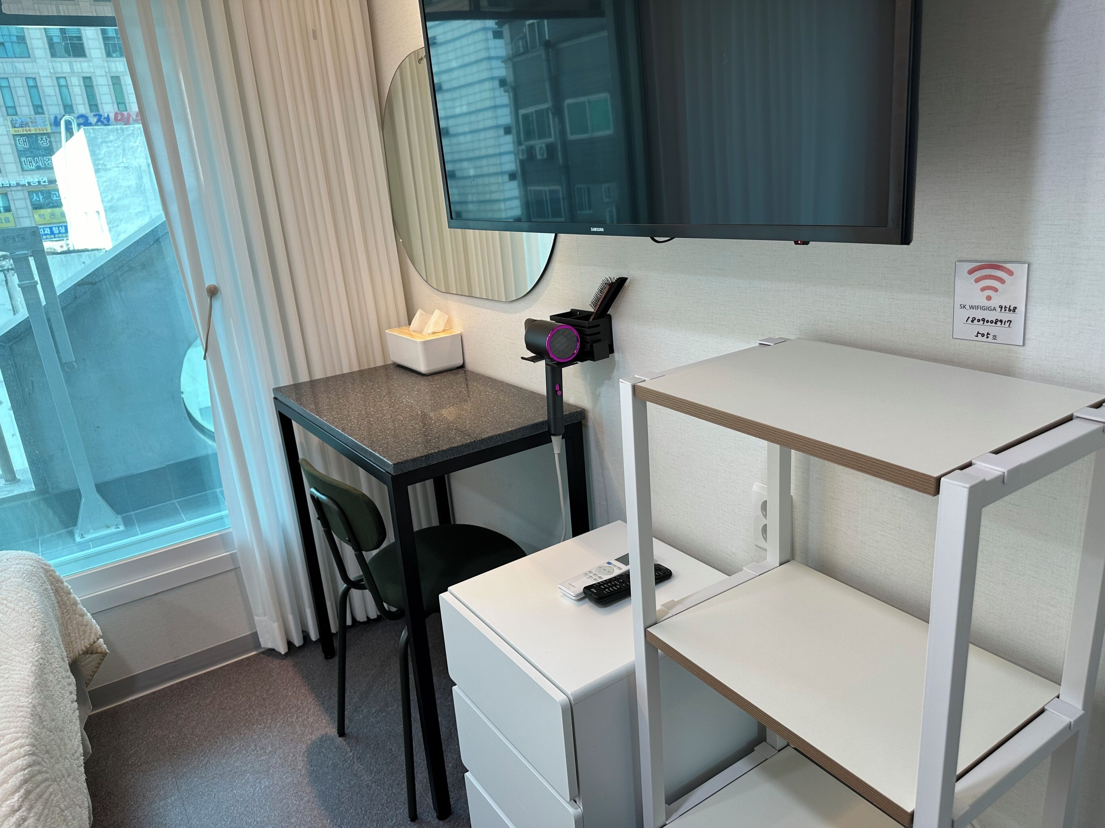 A hotel room with a bed, a desk with a chair, a TV mounted on the wall, a small table with a tissue box, a hairdryer, and a sign indicating WiFi availability.