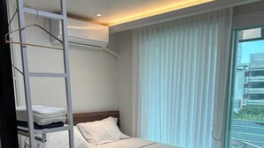 A hotel room with a bed, a white headboard, a window with curtains, a white wall, and a ceiling light.