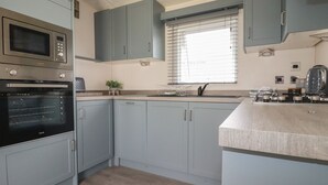A modern kitchen with grey cabinets, a built-in oven, a microwave, and a window with blinds. - No. 4 Fistral (Newquay)