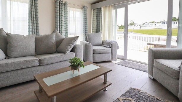 A modern living room with a grey sofa, a smaller armchair, and a wooden coffee table. There is a large glass door leading to a balcony with a view of a residential area. - No. 4 Fistral (Newquay)