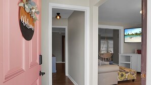 Et værelse med en lyserød dør, et 'Welcome' skilt, en hængestol, en sofa og et fjernsyn der viser et strandbillede.