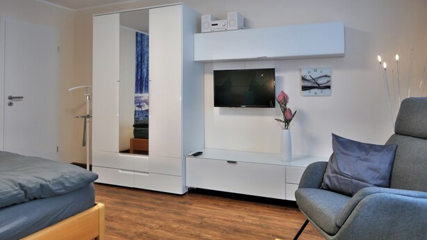 Een moderne kamer met een bed, een grijze fauteuil, een aan de muur bevestigde televisie en een kledingkast met spiegel.
