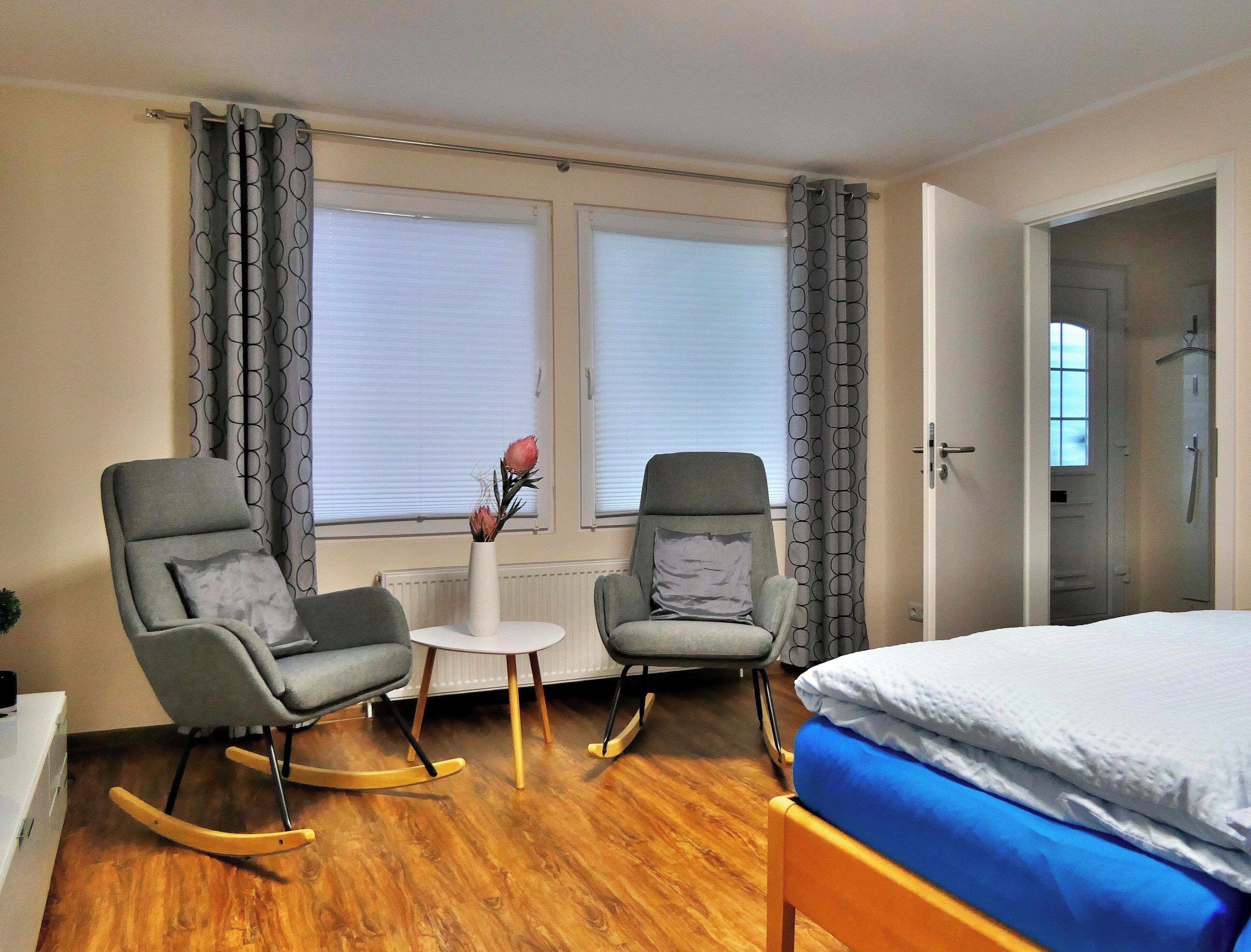 A room with two rocking chairs, a small table with a vase, and a bed with a blue mattress.