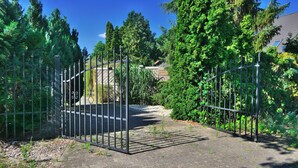 Een poort met een verhard pad dat door een aangelegde omgeving met bomen en struiken loopt.