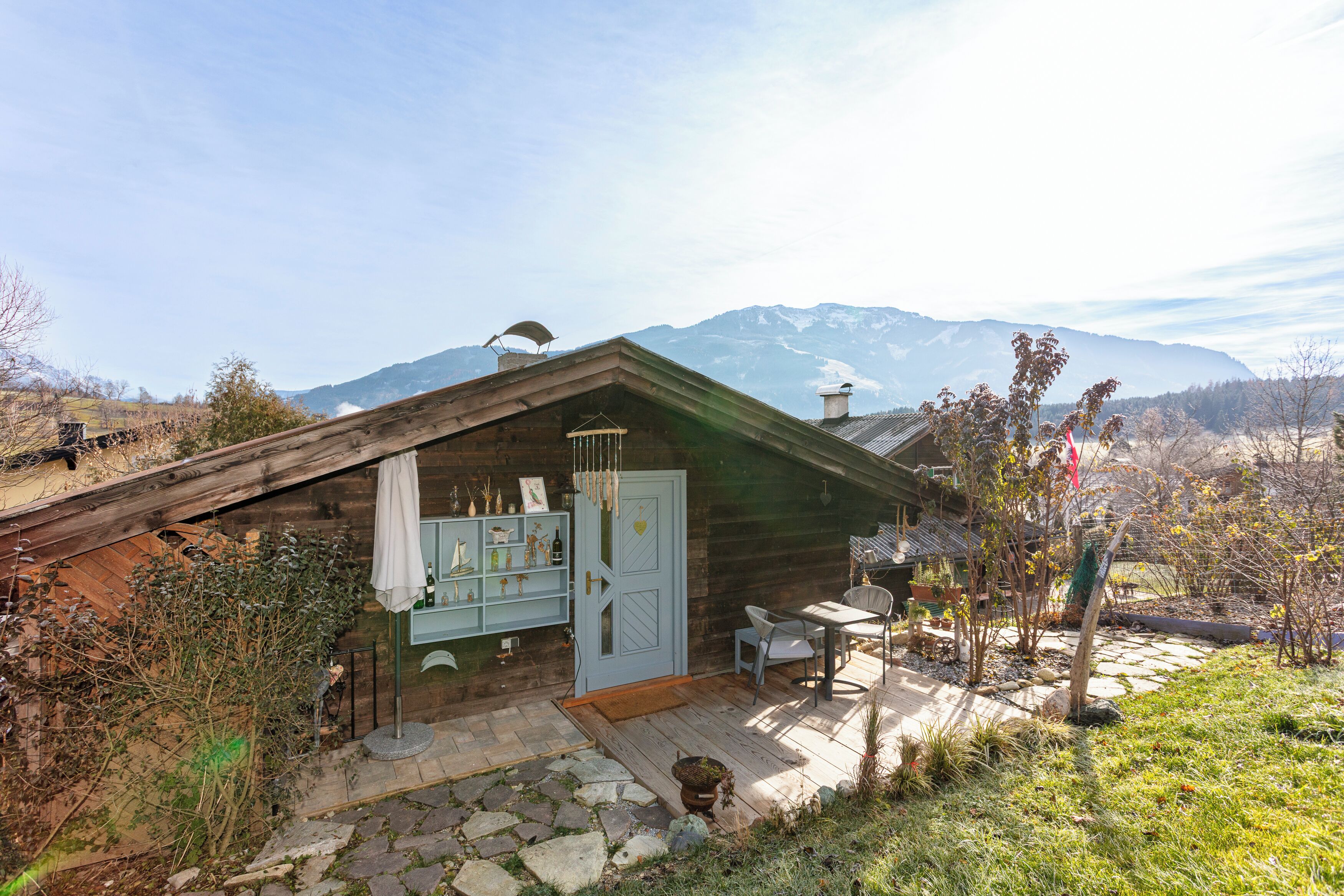 A wooden cabin with a stone patio, a small table and chair, and a view of mountains in the background.