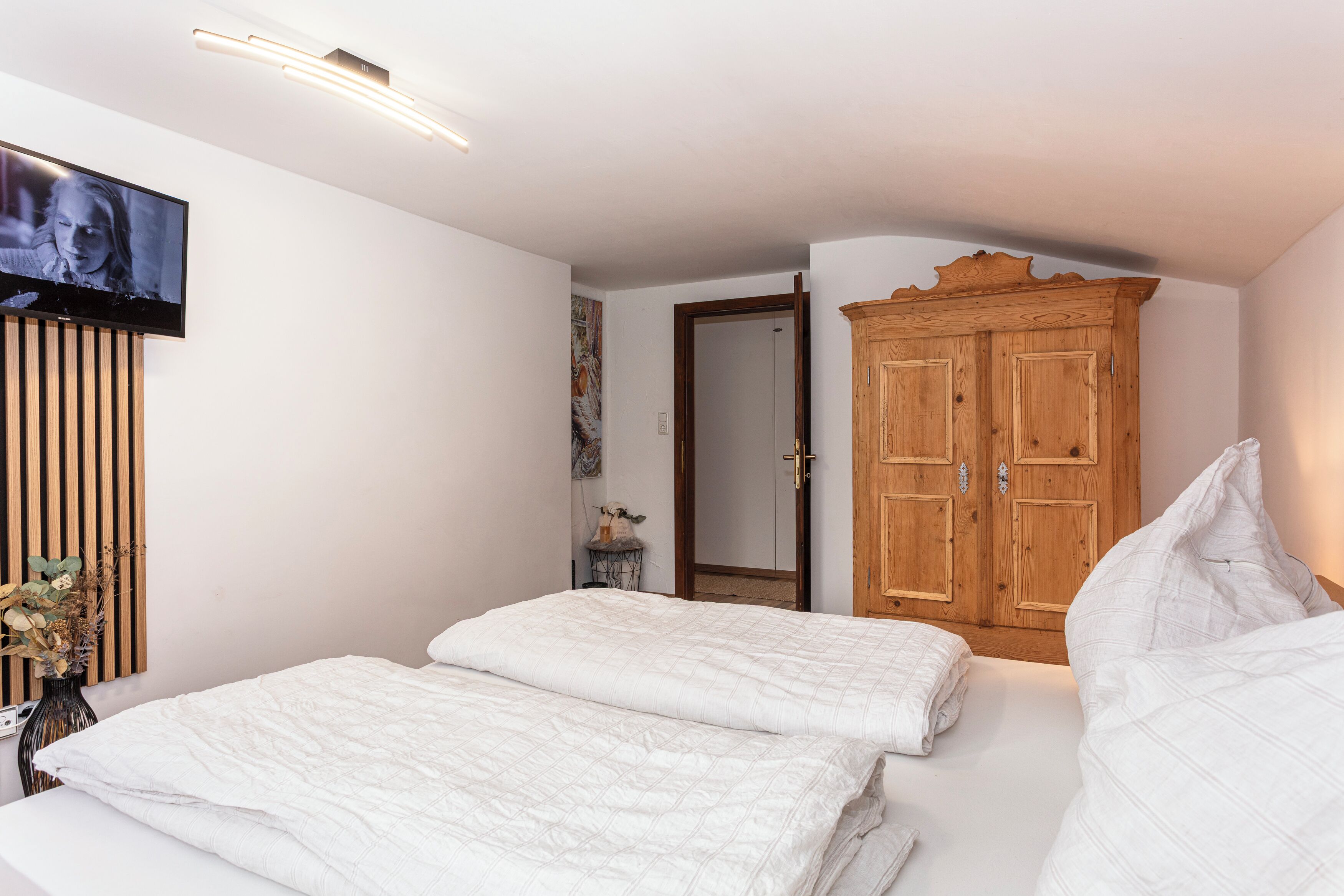 A bedroom with two beds, a wooden wardrobe, a television on the wall, and a vase with dried flowers.