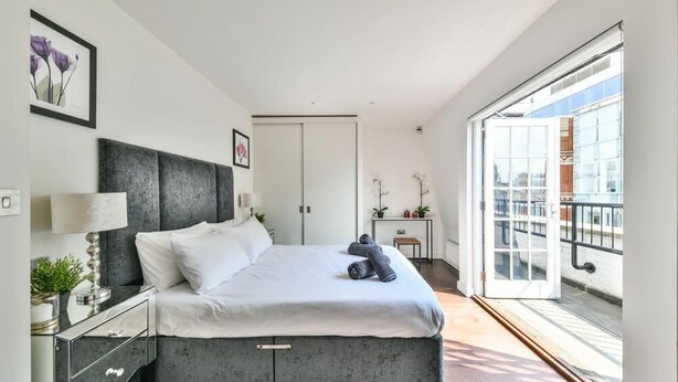 Un dormitorio moderno con cama, mesita de noche y vistas a la ciudad a través de amplias ventanas.