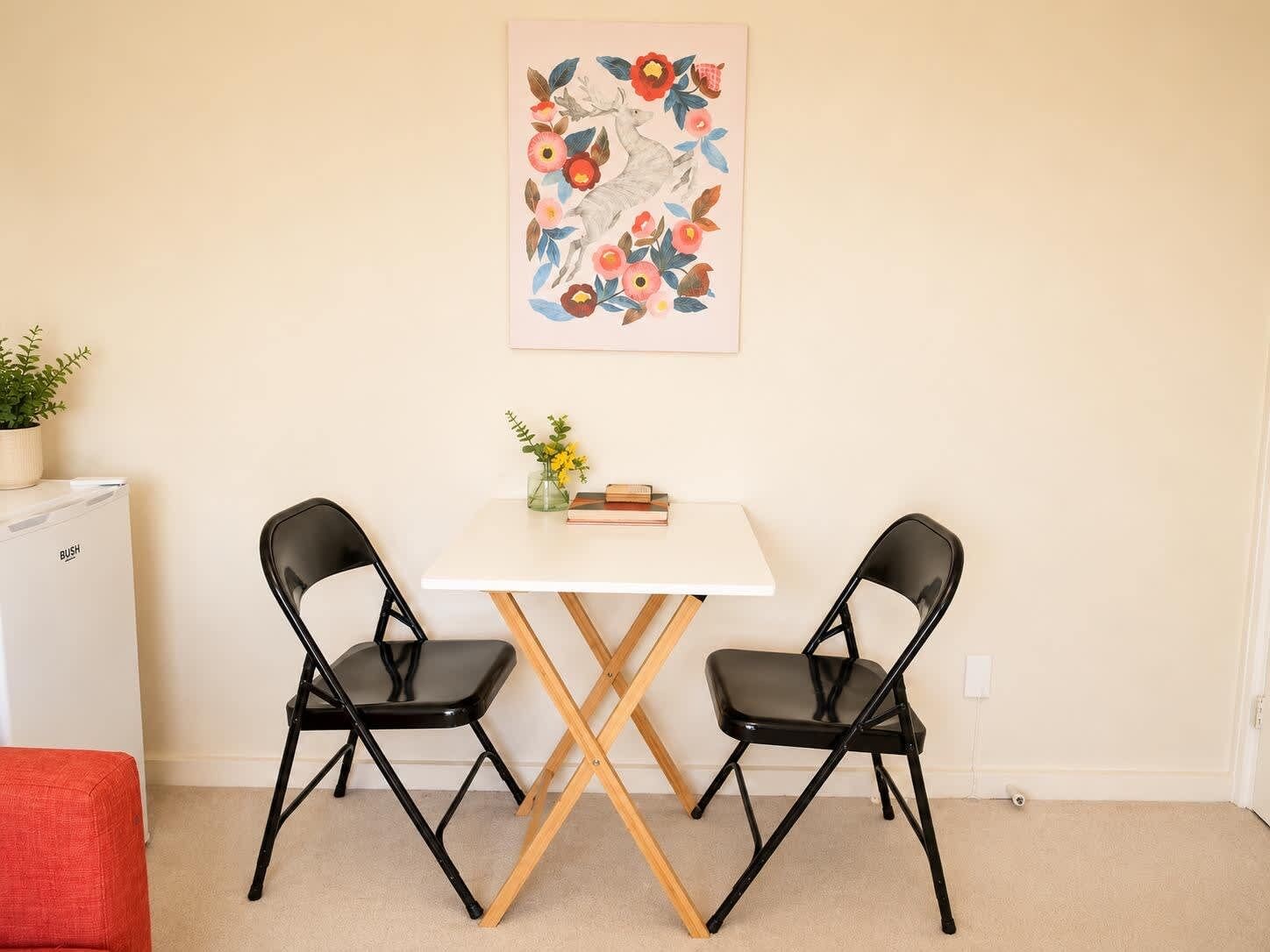 Een kamer met een witte tafel, twee zwarte stoelen, een kleine plant en een lijst met een foto aan de muur.