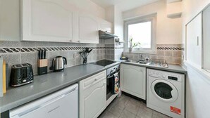 A modern kitchen with white cabinets, a built-in oven, a washing machine, and a window providing natural light. - The Elden - No.15 (Greater London)