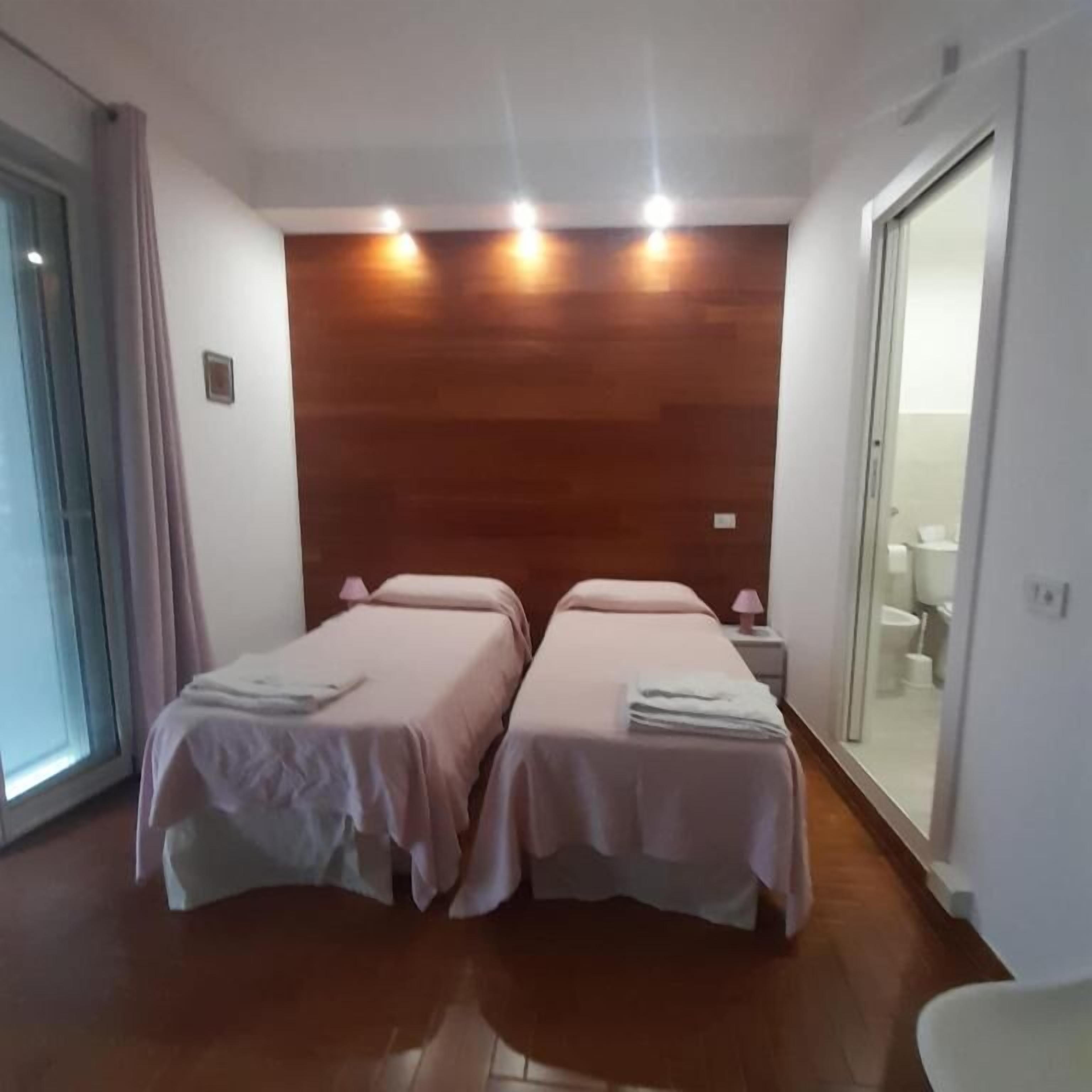 A hotel room with two beds, a wooden headboard, a small bedside table with a lamp, and a bathroom visible through a glass door.