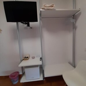 Una stanza con televisore montato su un supporto, una sedia bianca, uno scaffale con un asciugamano arrotolato, un piccolo tavolino con un telecomando e un cestino.