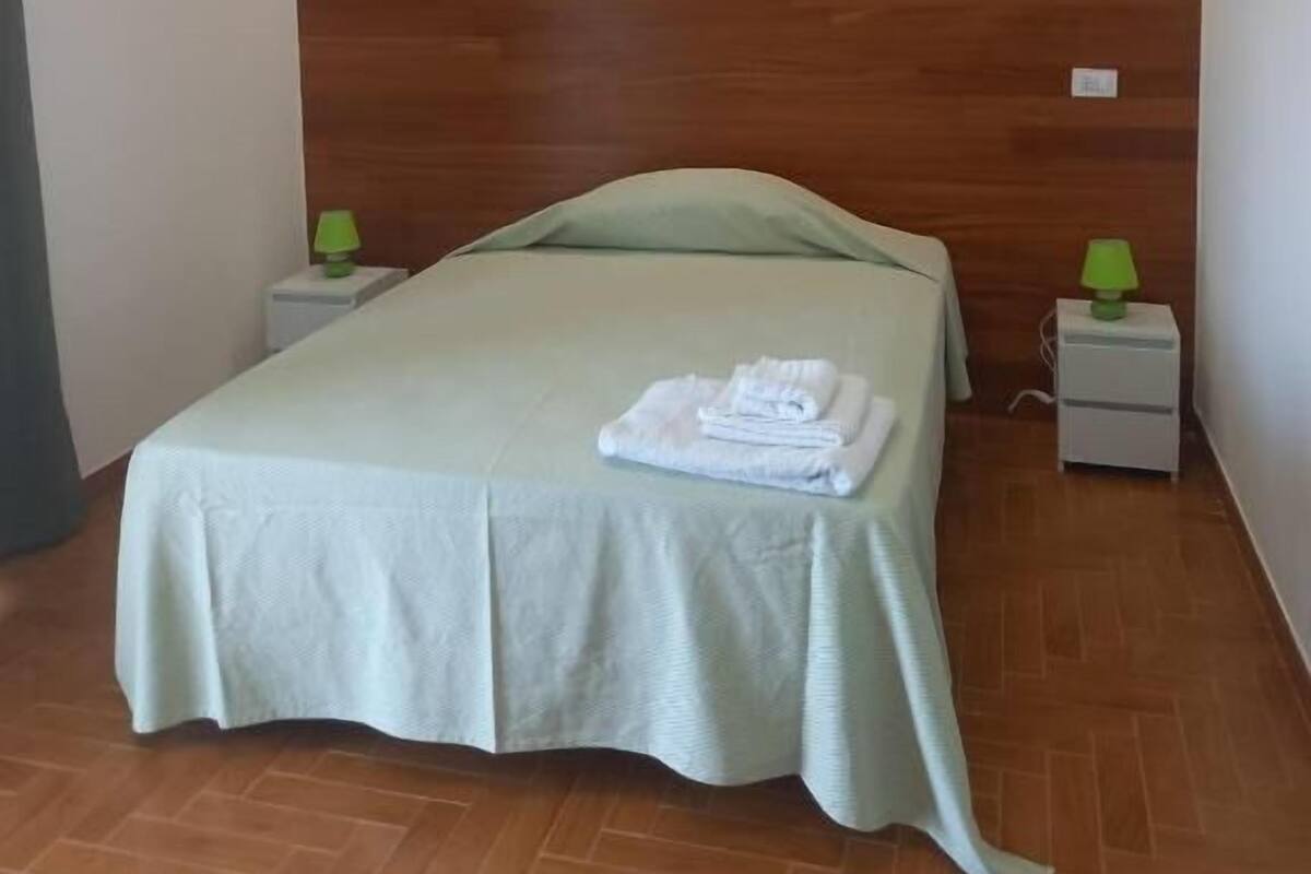Makuuhuoneessa on puinen sängynpääty, vihreällä päiväpeitteellä varustettu sänky, kaksi yöpöytää, joissa on lamput, ja taitettu pyyhe sängyllä.
