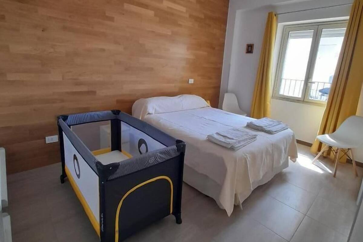 Hotellihuone, jossa on puinen seinä, valkoisilla lakanoilla varustettu sänky, pieni leikkiaita ja ikkuna, jossa on verhot.
