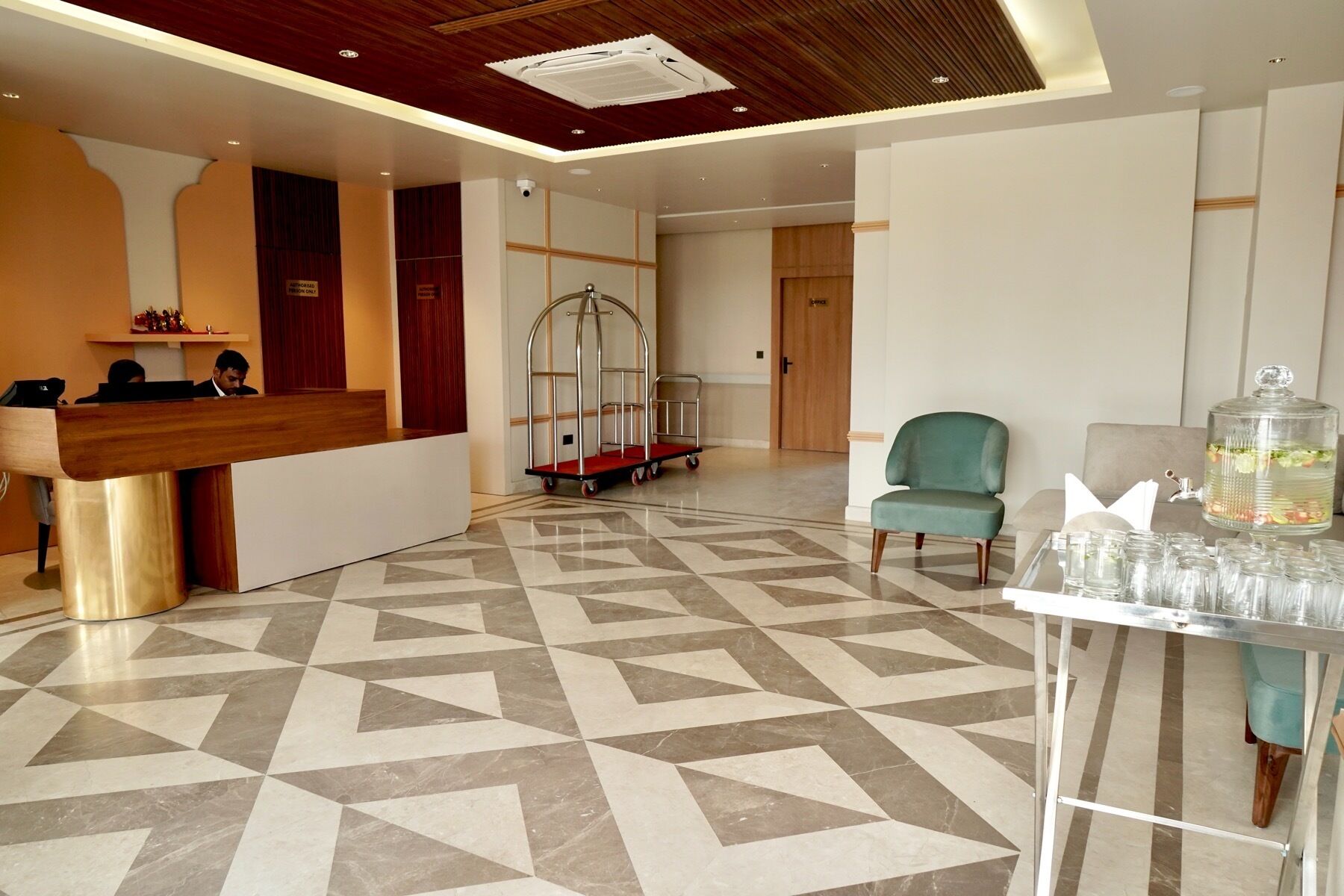 Un vestíbulo de hotel con una recepción, un área de espera con una silla verde, un carrito y una mesa con vasos y dispensador de agua.