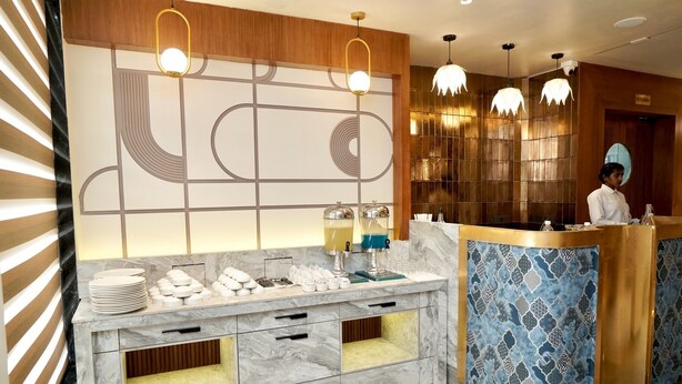 Uma área de recepção de hotel moderna, com balcão de mármore, dois dispensadores de bebidas e uma parede decorativa com padrões geométricos.