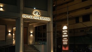 Exterior do Hotel HPB Grand com sinalização iluminada, uma escadaria que leva à entrada e uma área de estacionamento com motocicletas.