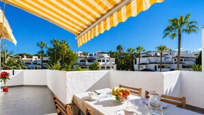 A table set outdoors with a yellow striped umbrella, a white tablecloth, and a view of white buildings and palm trees. - Penthouse on the beach - Elviria (marbella)
