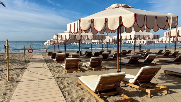 A beach with wooden lounge chairs and white umbrellas with red trim, arranged on a sandy area with a wooden walkway. - Penthouse on the beach - Elviria (marbella)